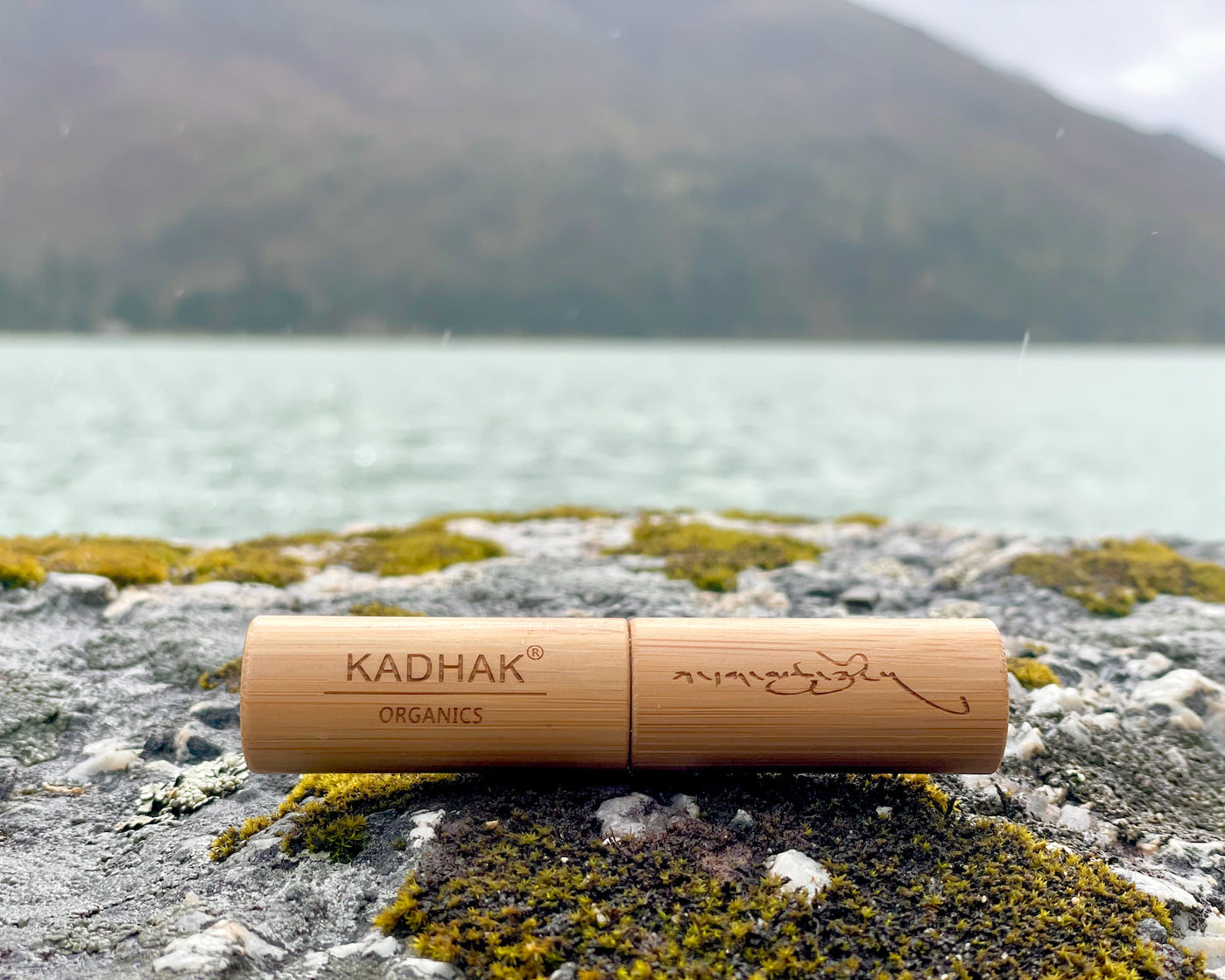 Bamboo container with 'Kadhak Organics' branding on a rocky surface with a lake and mountains in the background